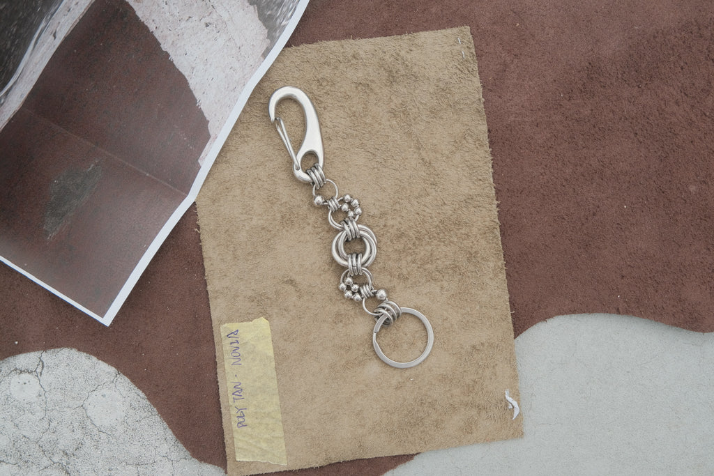 A silver metal keychain with a lobster clasp, decorative chain links, and a split key ring, displayed on a piece of brown paper over a textured brown and white surface.