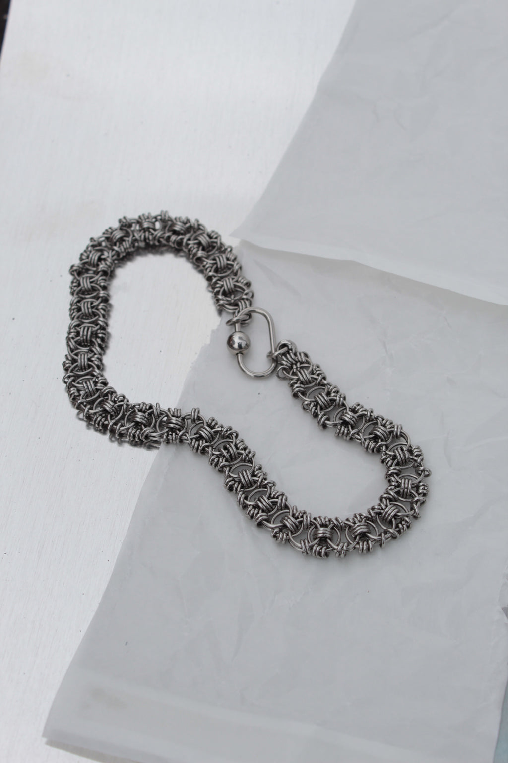 A silver-toned chainmail-style bracelet with a spring-ring clasp, displayed on a white textured surface.